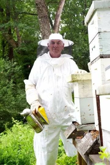 Tom Curren wearing his beekeeping attire and standing in front of bee hives.