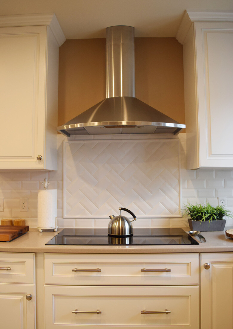 Elegant kitchen remodel in Waltham, MA, by Tom Curren, featuring a stainless steel range hood, white shaker cabinets, and a herringbone tile backsplash.jpg